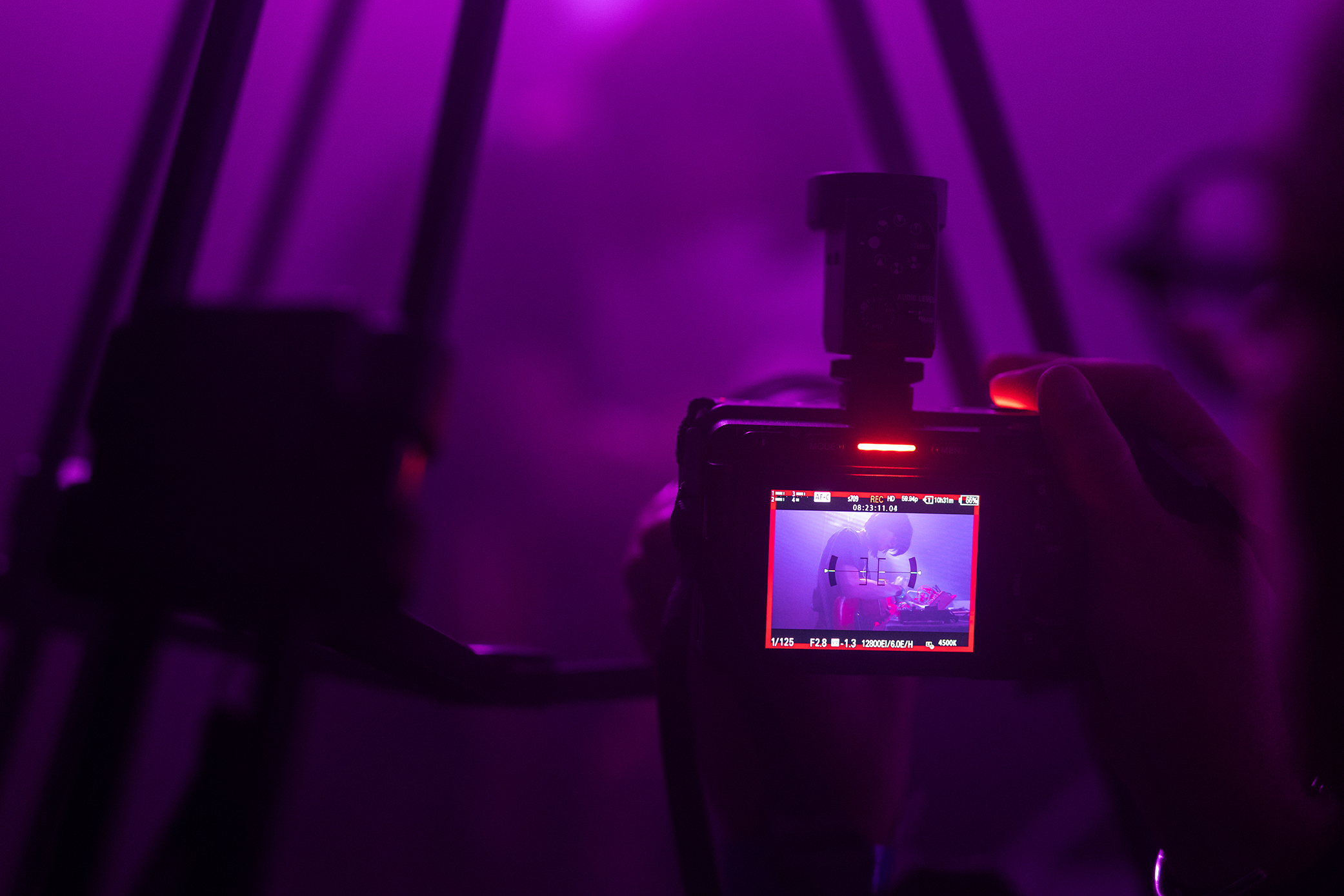 A camera viewfinder shooting a live act on stage, purple stage lighting fills the shot