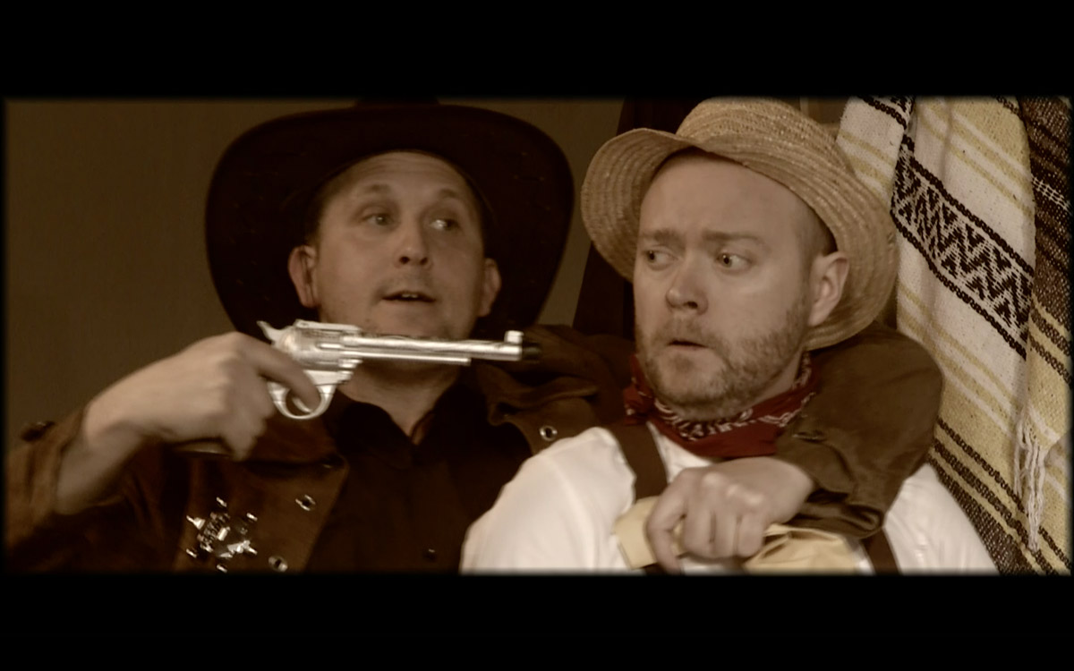 Man in cowboy-style hat holds a gun to the head of a man.