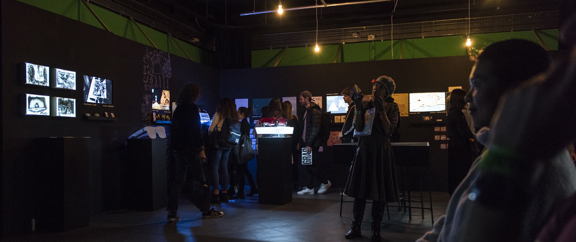 Exhibition shot of a dark space with visitors watching screens and interacting.