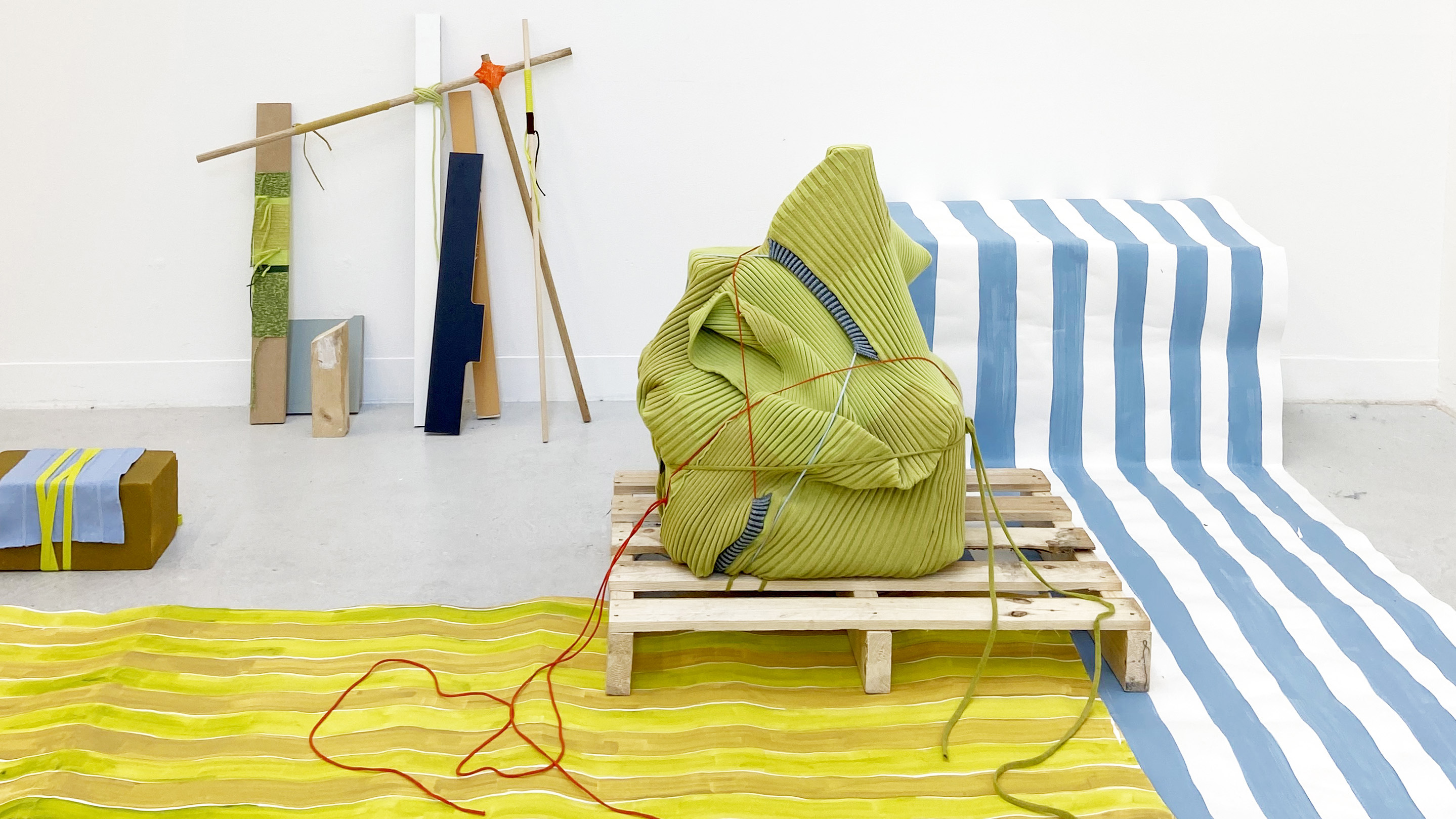 A room shot of colourful fabrics laid across the floor. There is a bean bag structure wrapped in red string sitting on a pallet