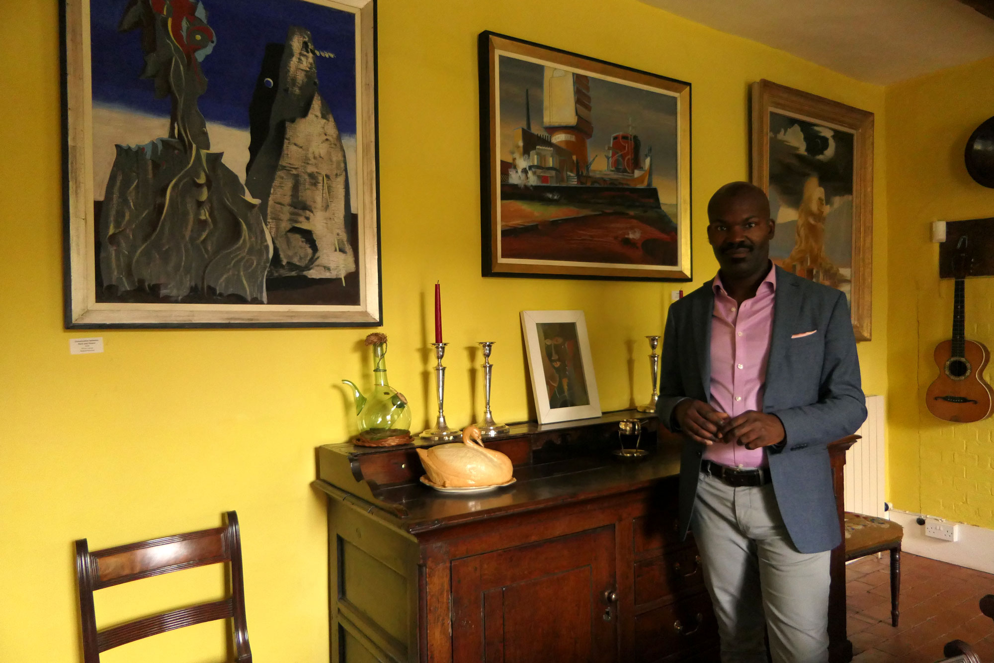 A man stands in a yellow room hung with framed works of art.