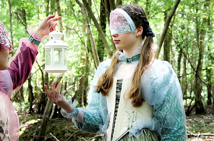 Two masked models interact with eachother while in a forest looking away from the camera wearing garments inspired by the pre-Raphaelite art movement