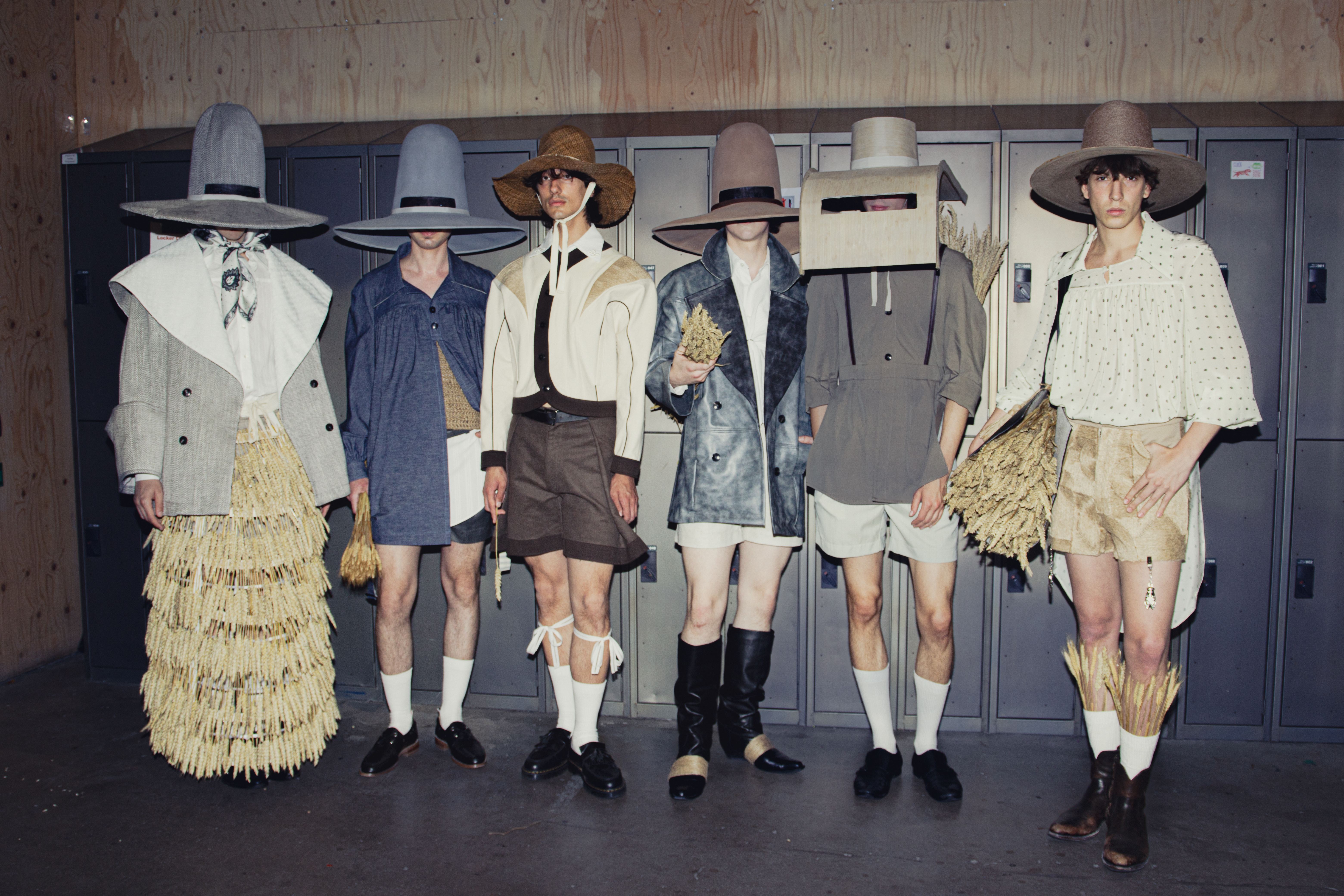 A menswear collection on a row of 6 models standing in front of aluminium lockers. The clothes reference nature and the countryside with straw or crop-based embellishments, three quarter socks and brimmed hats