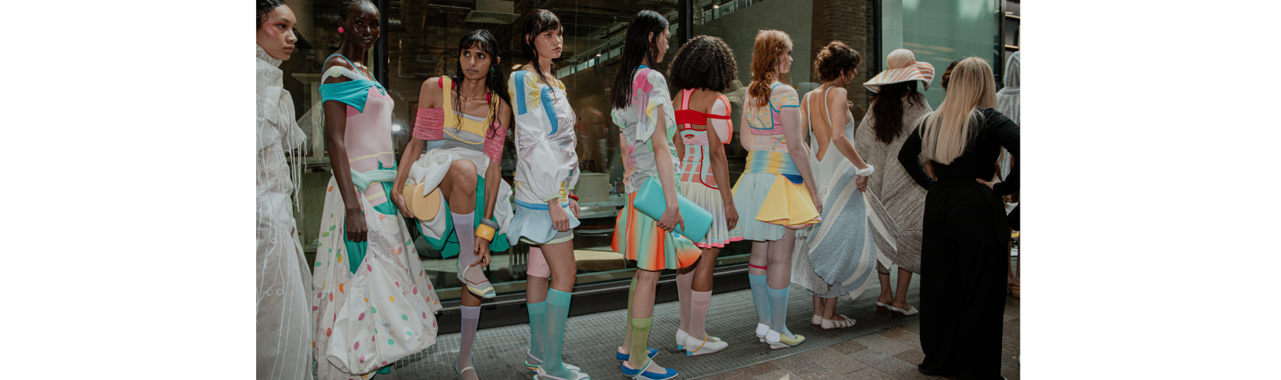 Shot from the side, a long row of models in colourful sheer and knitted garments referencing retro sportswear are lined up ready to go on the runway.
