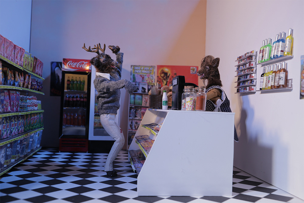 A male model of a deer, taken as a still from a stop motion animated film, waves to another deer in a shop scene.