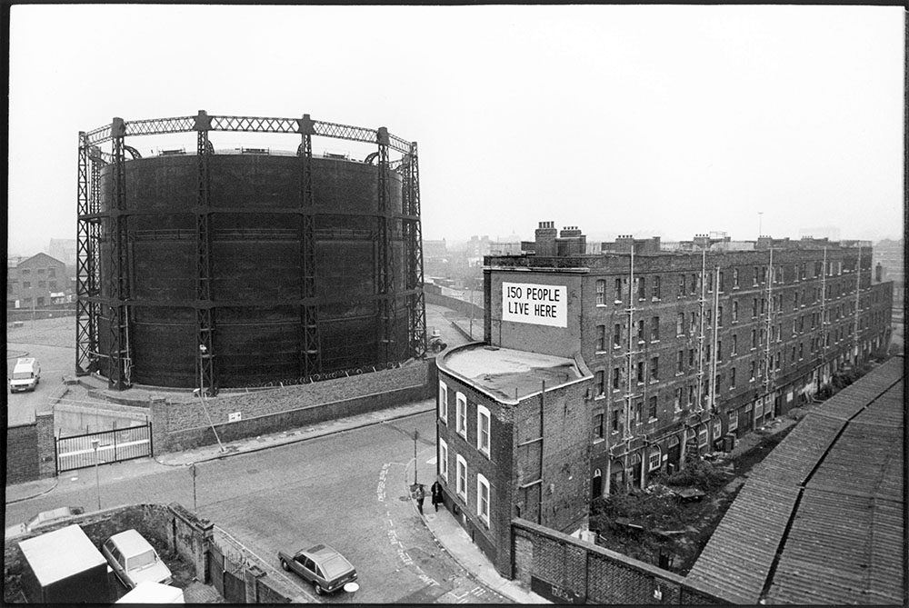 Black and white photo of a gasometer with nearby building written '150 people live here' written on it