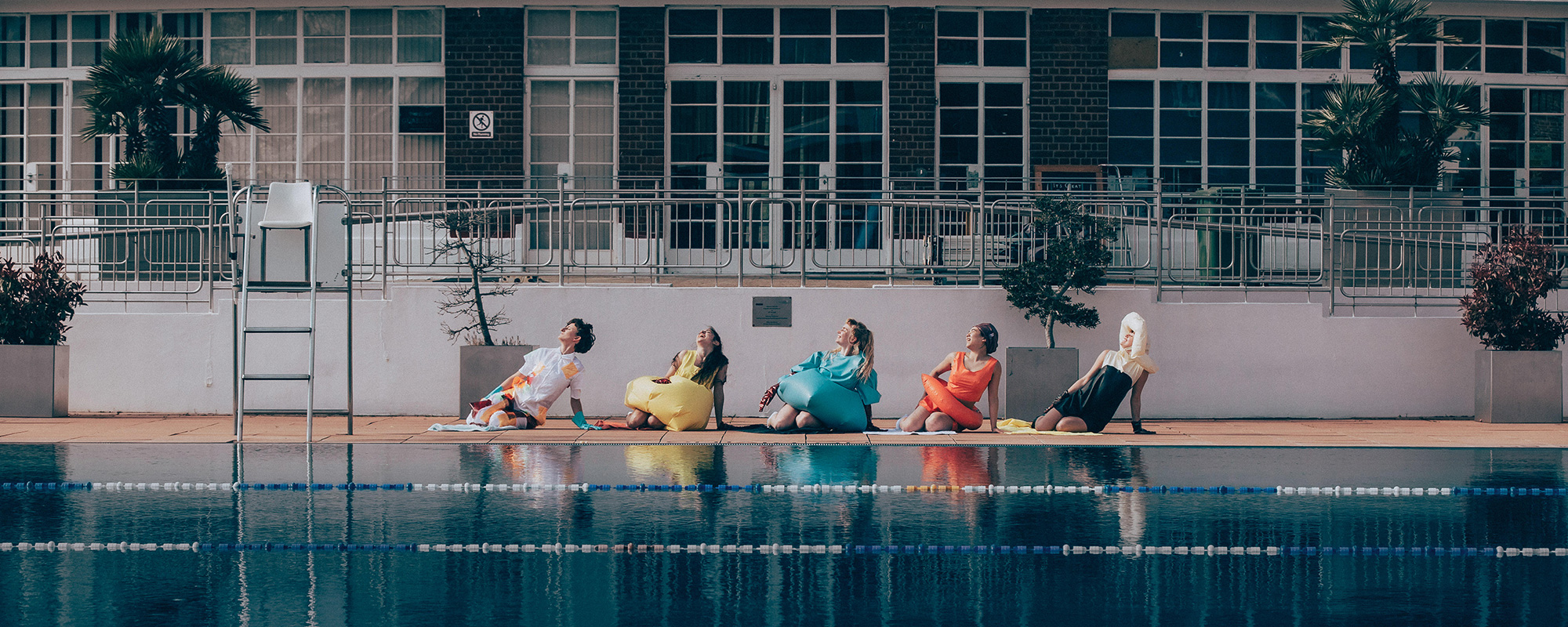5 adults pose in brightly coloured outfits by an outdoor swimming pool