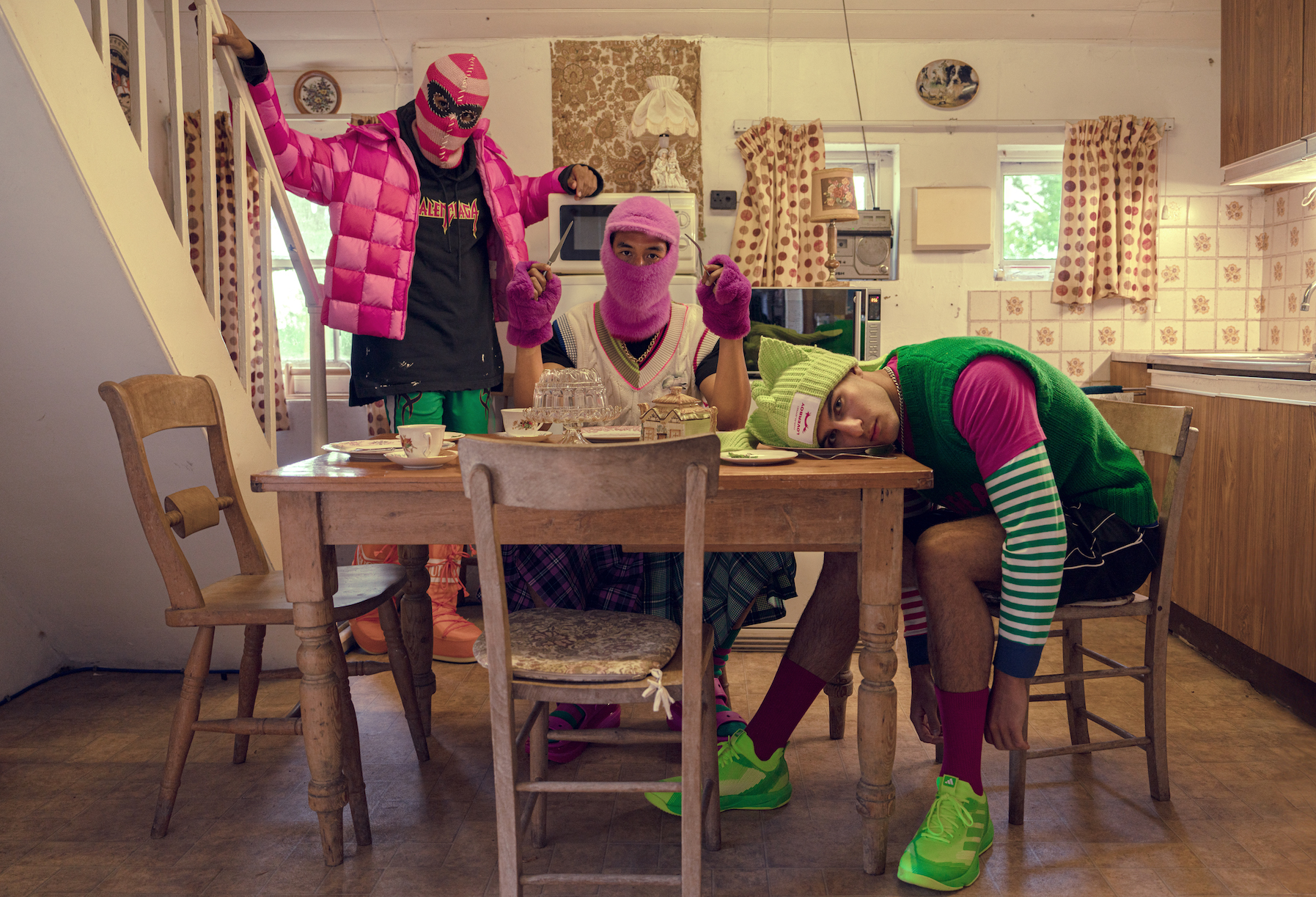 A stylised editorial shot of 3 people at a kitchen table, they are wearing neon hats and balaclavas and one has their head on the table while the others pose with arms up