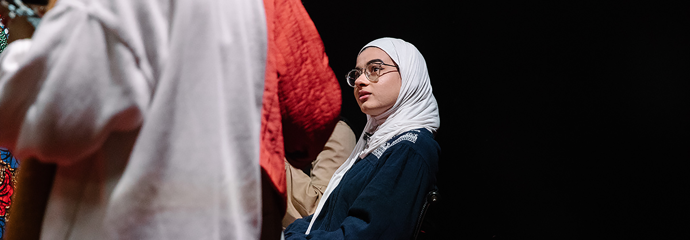 Woman in hijab with glasses listening in crowd in dark room