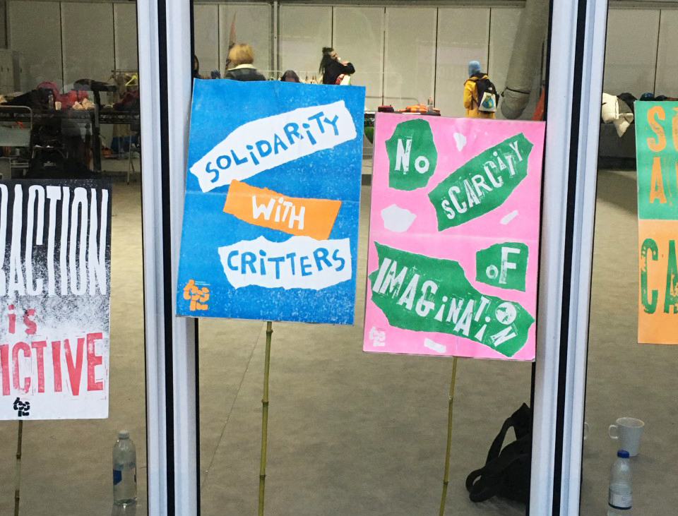 4 placards lay against a window. One is blue and white and says, 'solidarity with critters' and the other is pink and green and says 'no scarcity of imagination'.