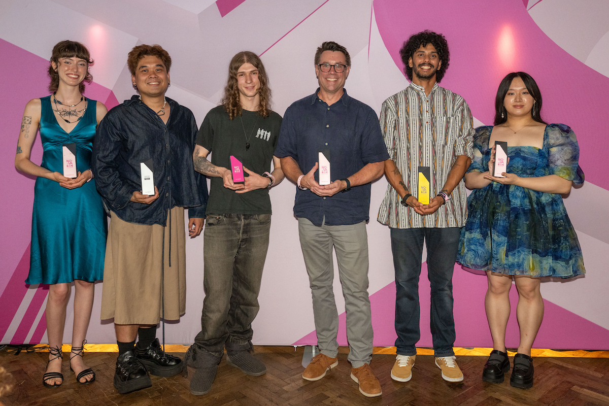 A group of student award winners stand in front of a pink background holding their awards