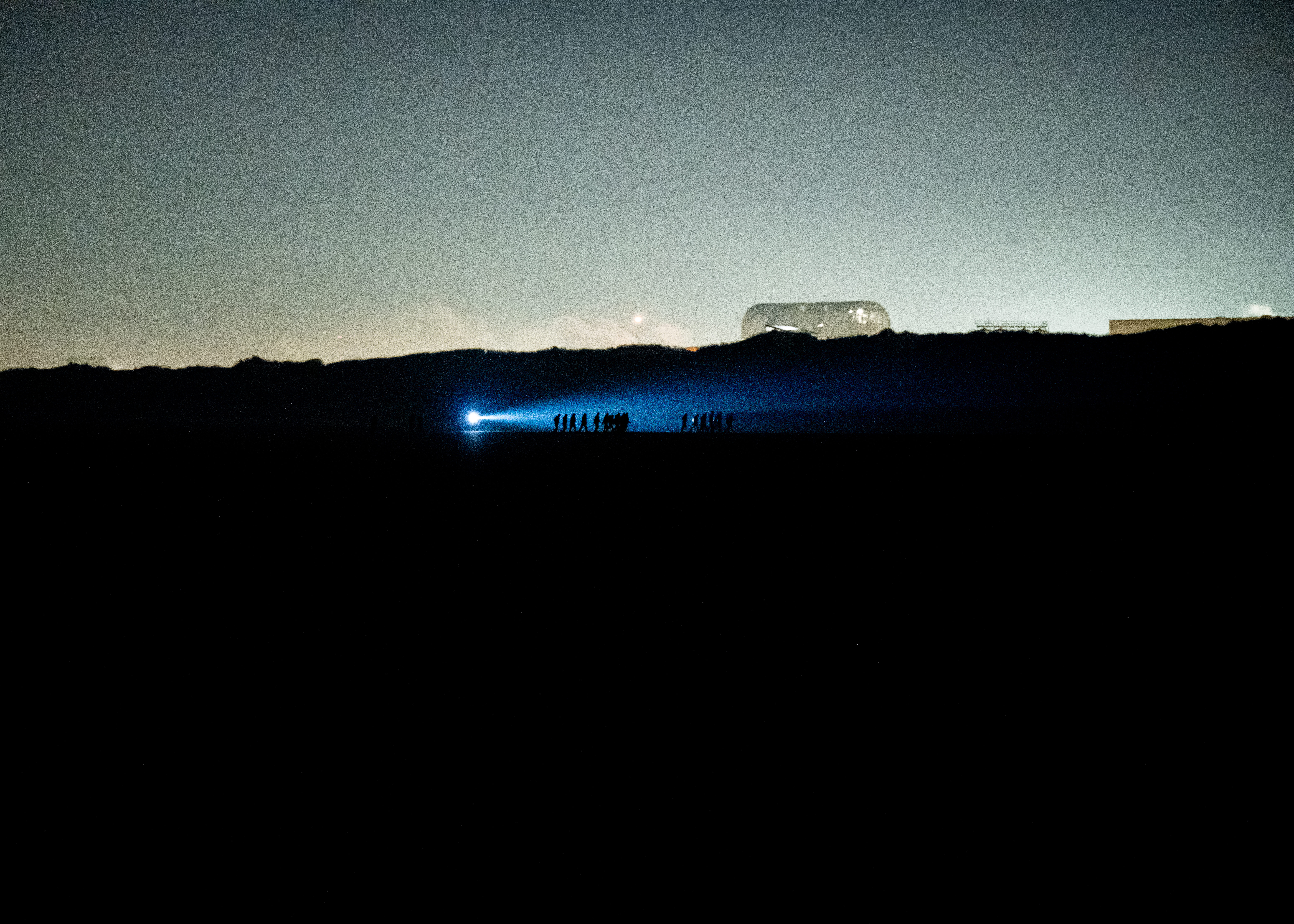 A photo of a landscape at dusk with human figures walking in silhouette, next to water under a strong outdoor torchlight 