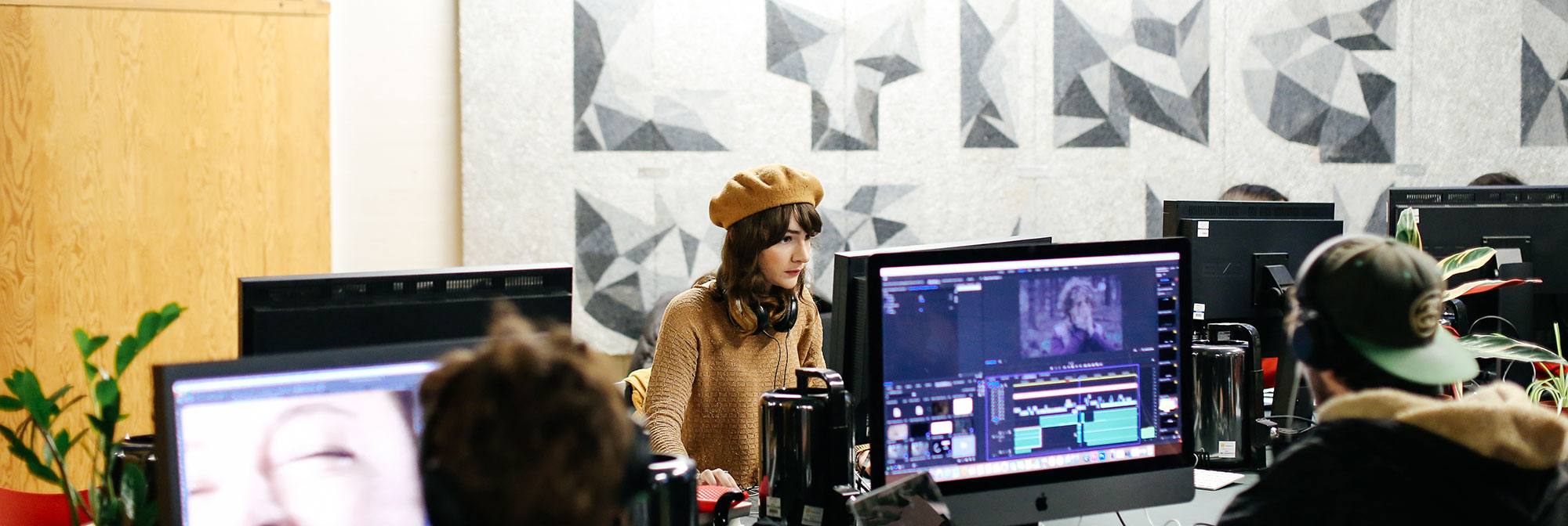 A woman in a beige beret sits at a computer screen. A man in a black cap sits with his back to the camera so that we can see his screen which shows purple and blue graphics.
