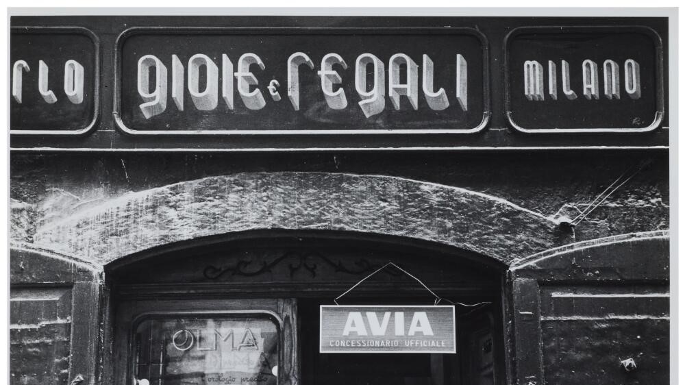 Black and white photograph of a shop sign with an elongated letter style, reading, ‘Gioie e Regali, Milano’.