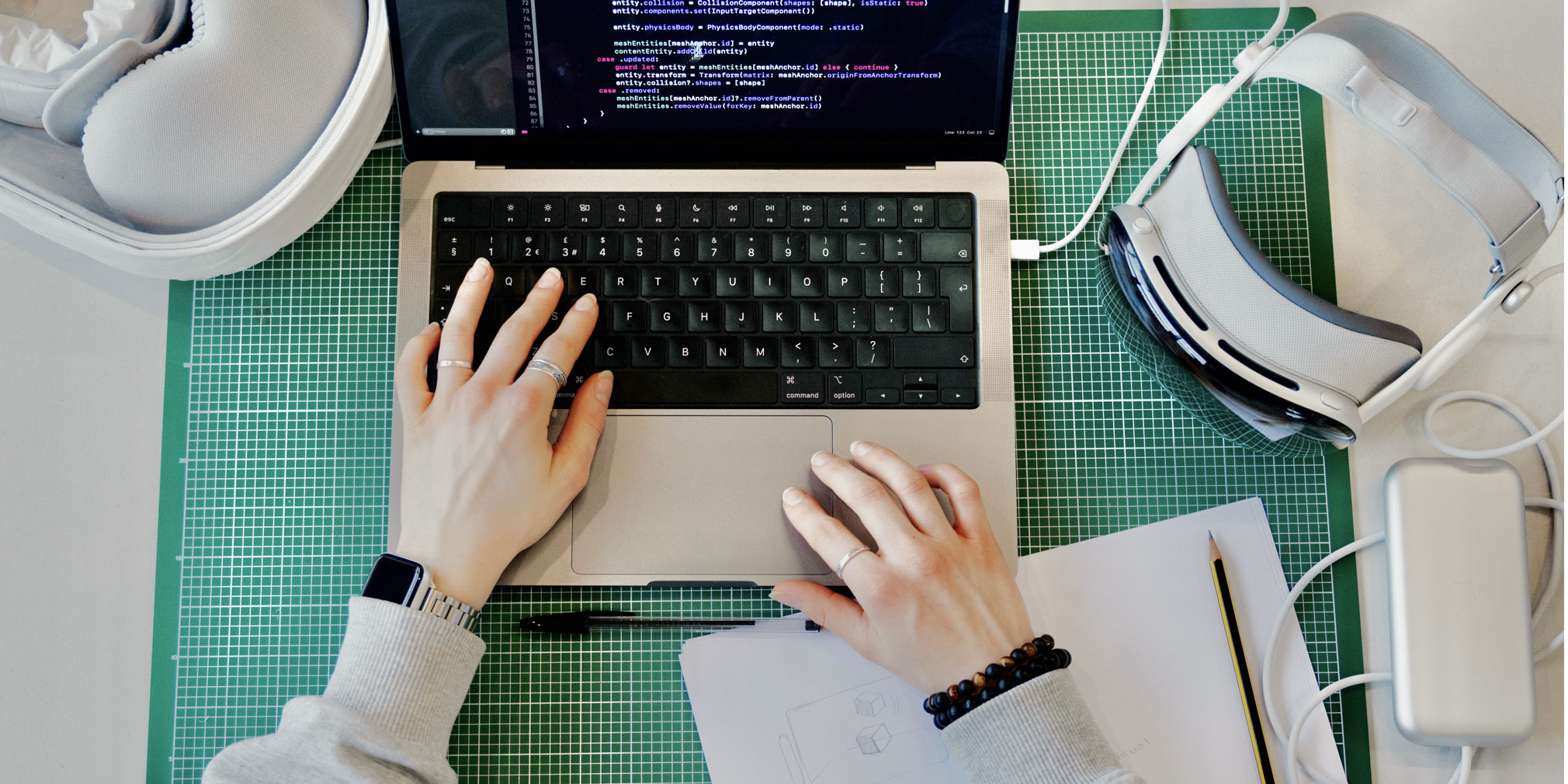 MacBook Pro with colourful Swift code displayed, and an Apple Vision Pro off to the side, on a green cutting mat, with hands on the keyboard.