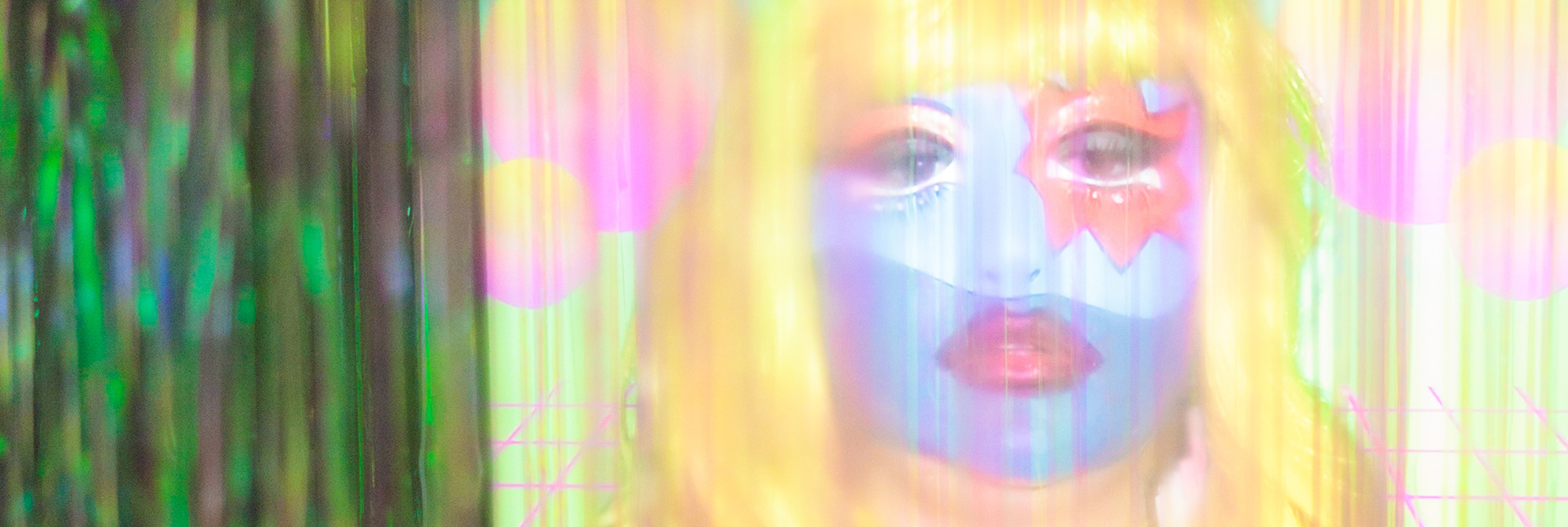 Close-up photograph of a screen showing a yellow and pink video of a woman in blue and red face make-up, wearing a yellow wig, is viewed through a curtain of iridescent green plastic streamers.