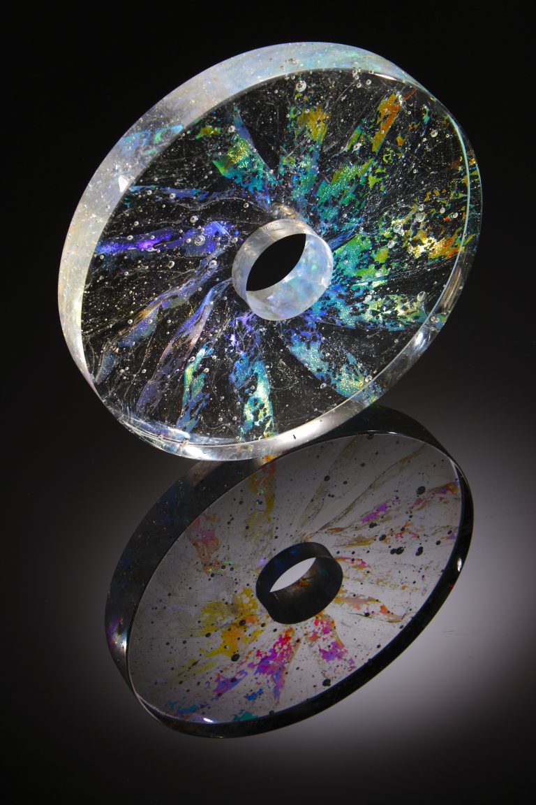 Glass circle with hole in the middle. Transparent glass with multi-coloured metalic slices radiating from the middle towards the edges and bubbles trapped inside.