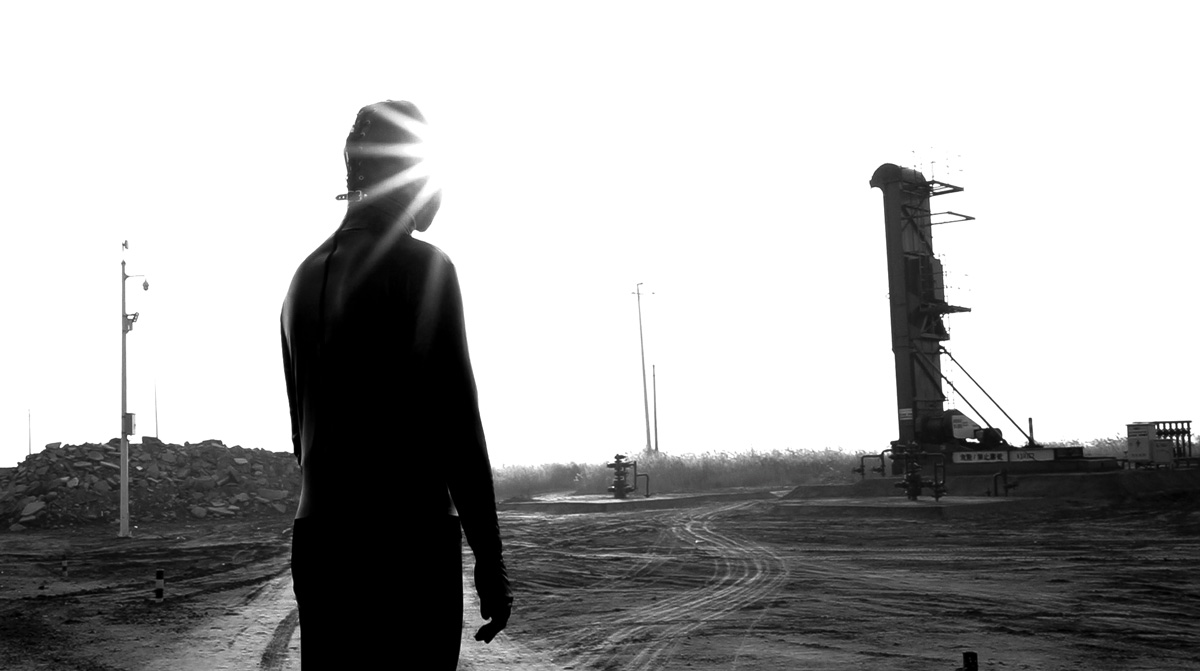 Person in a leather mask stands in front a industrial site in this black and white photo
