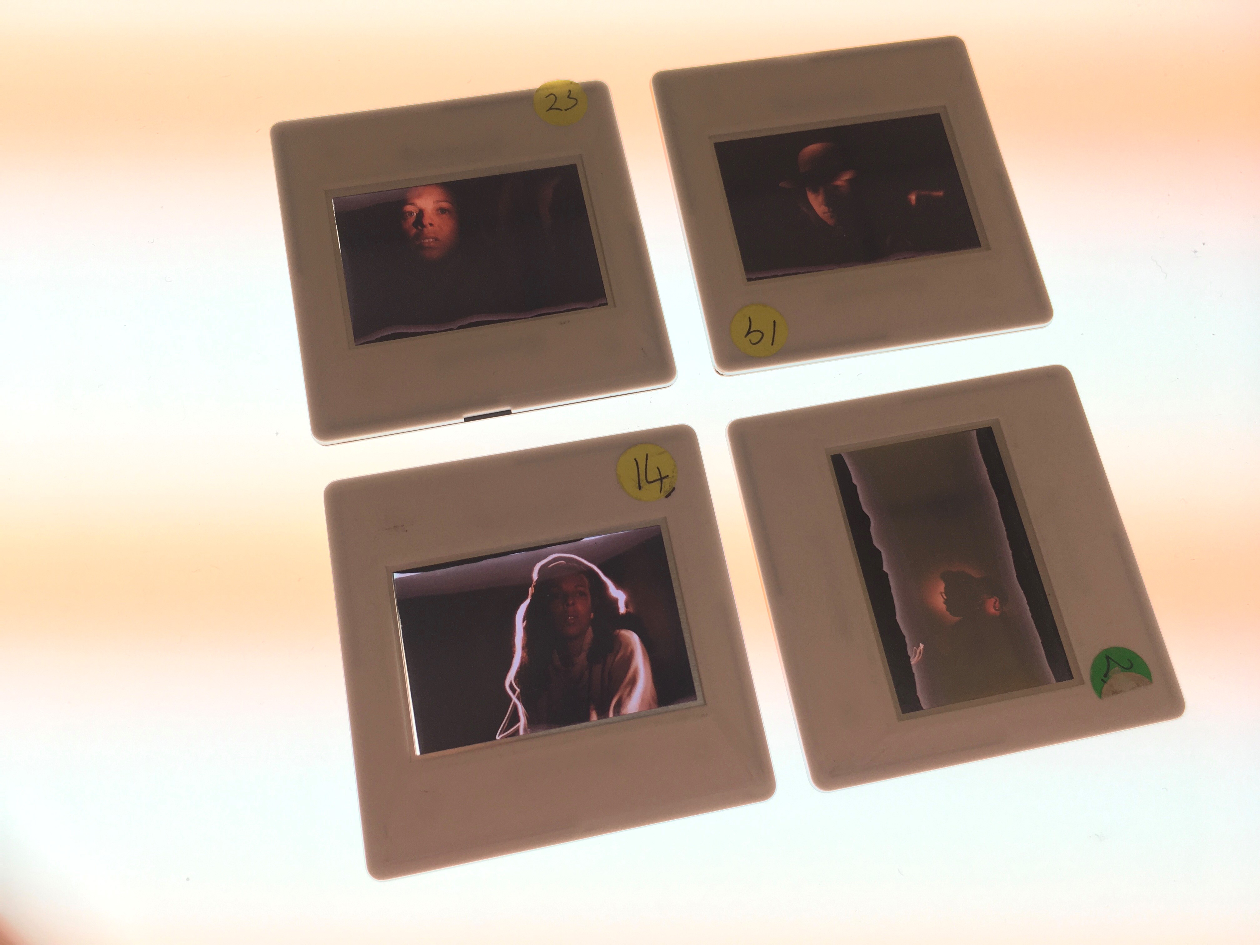 Colour image of 4 slides lit from the back, resting on a light box. 35mm colour transparencies mounted in standard white plastic slide mounts, labelled on the corners with round yellow stickers with numbers on them. The images appear to show portraits of women of colour