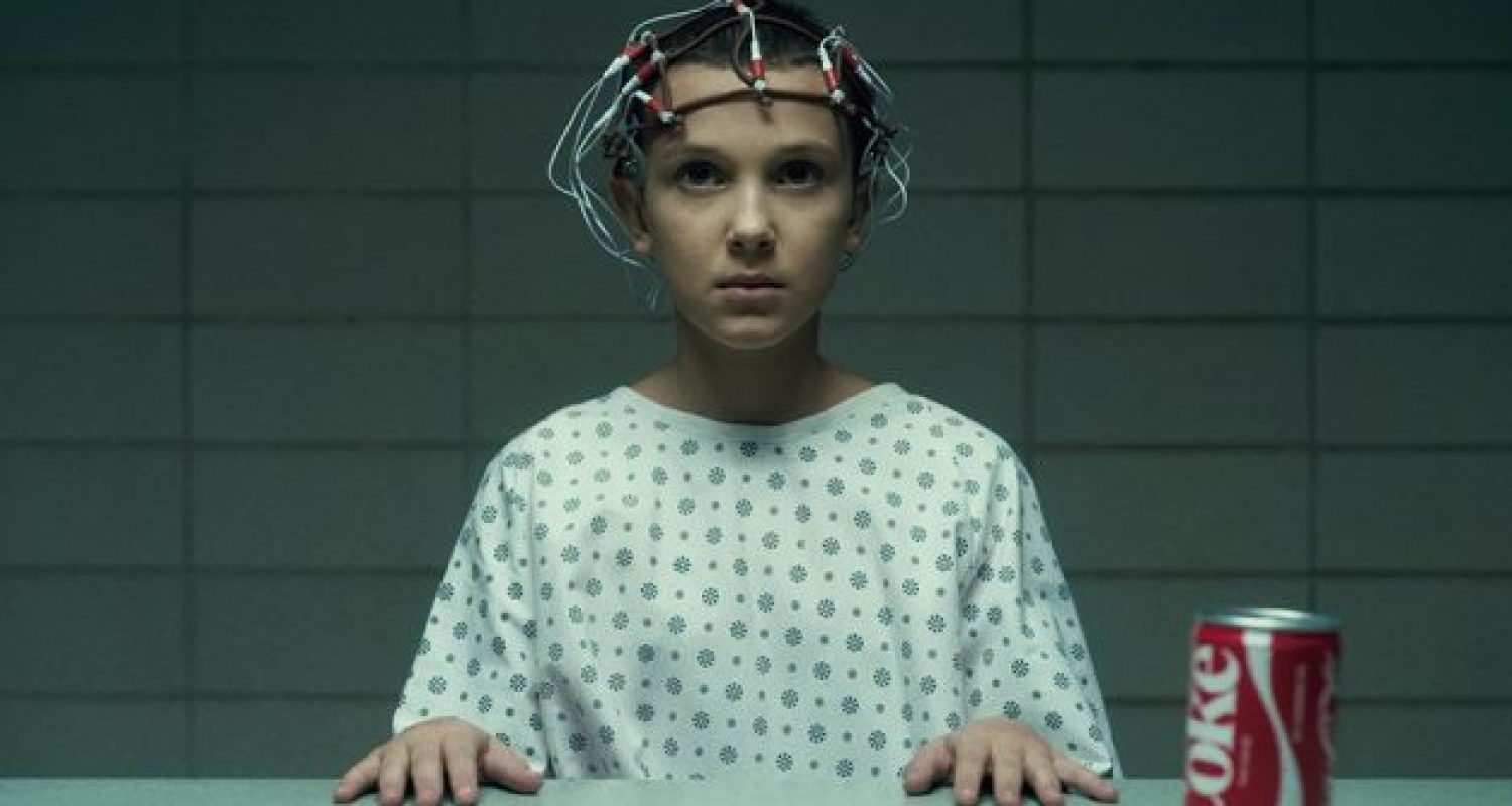 Still from the television programme Stranger Things, showing Eleven sat with an electrode helmet by a table with a Coke can.