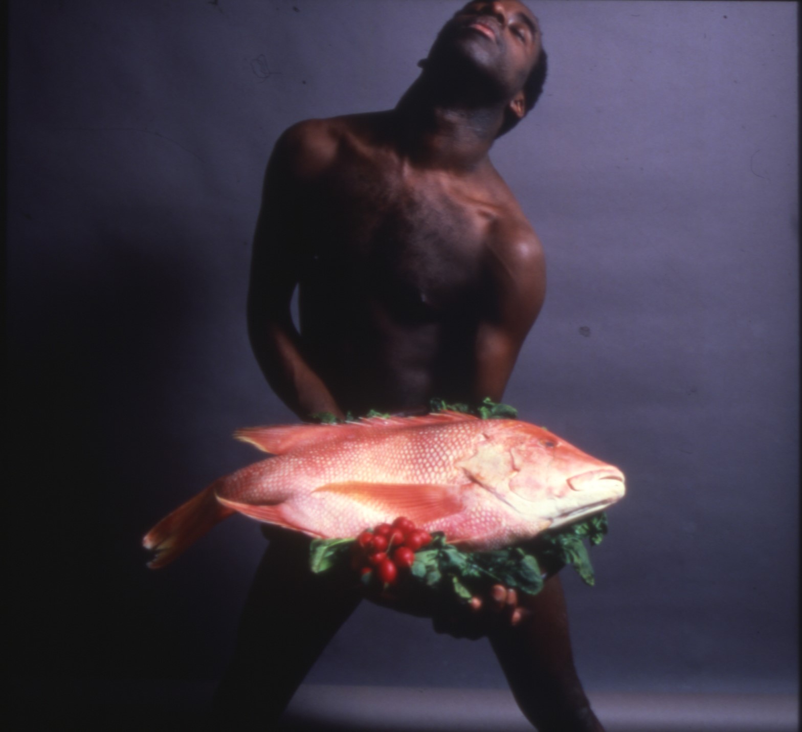Colour photograph of a young black man, nude, with his eyes closed, holding a large fish, possibly a red snapper