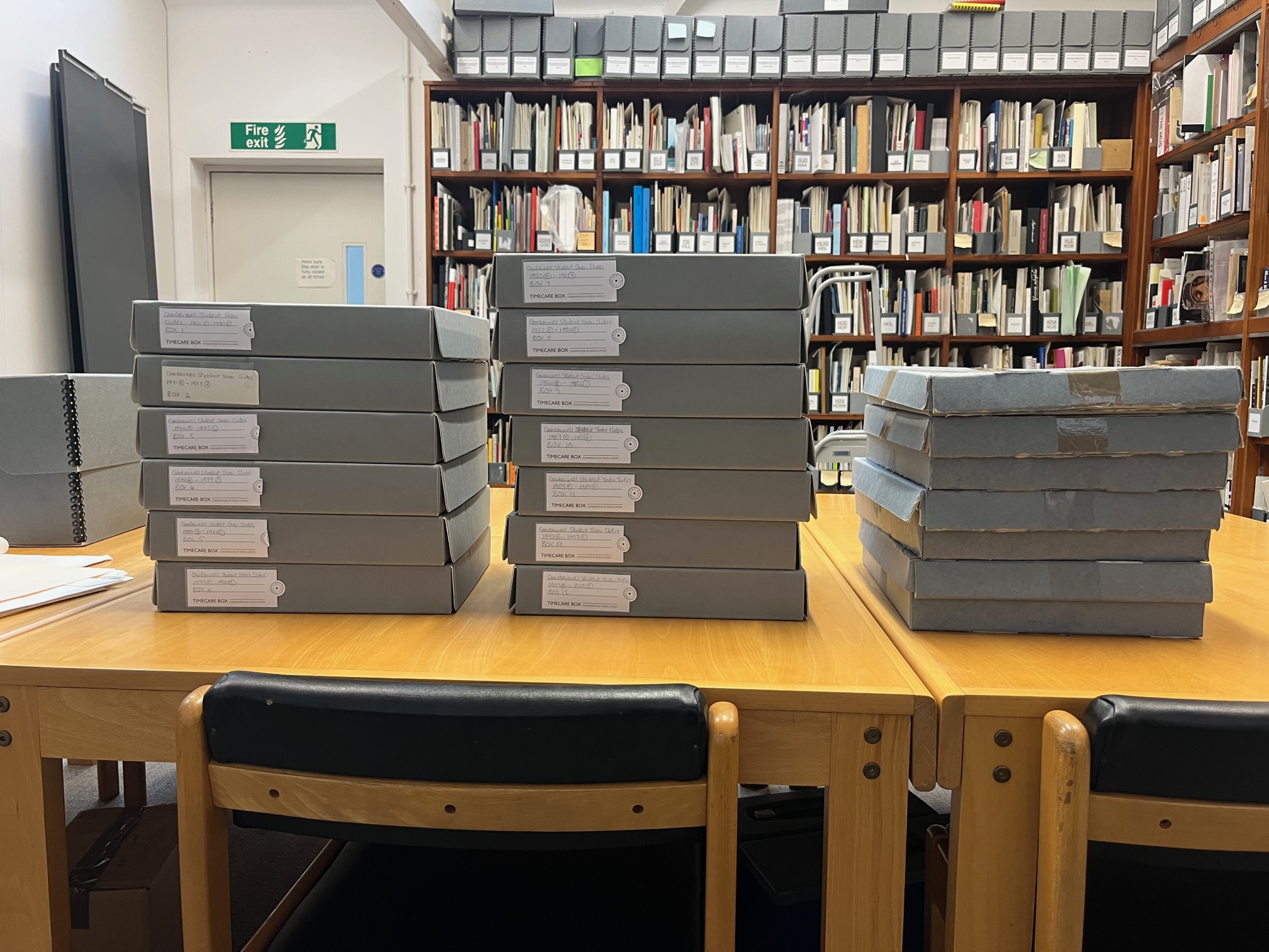 Colour photograph showing a set of archival boxes on a table in front of shelves with archive and library material on them 