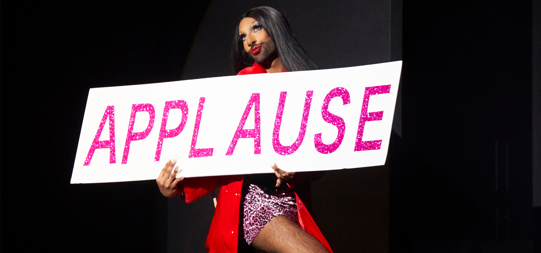Bearded, long haired man in pink boxer short and lipstick holding a large sign stating APPLAUSE