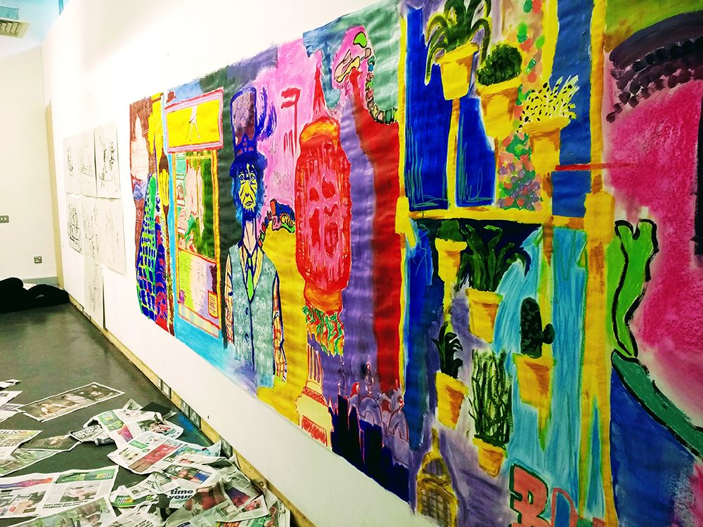 A large vibrantly coloured painting pinned to the wall featuring a man in a tall hat, potted plants, buildings and shop fronts.