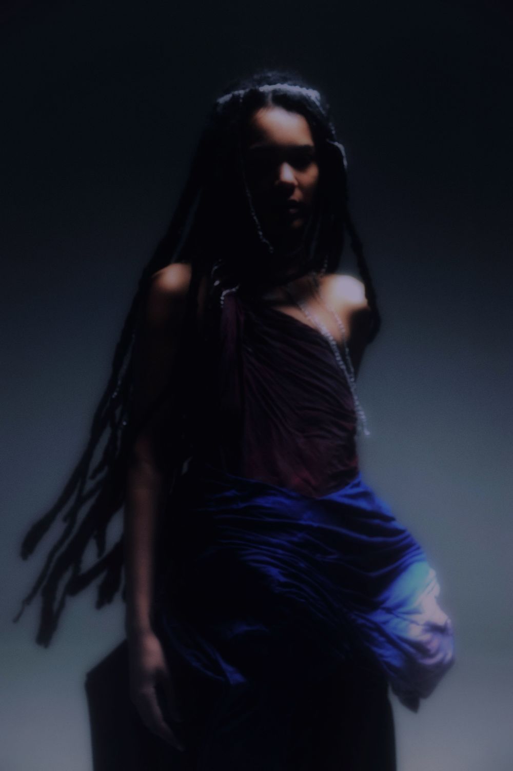 Figure wearing dark dress, with long hair and crystal necklaces, set against a vignetted background