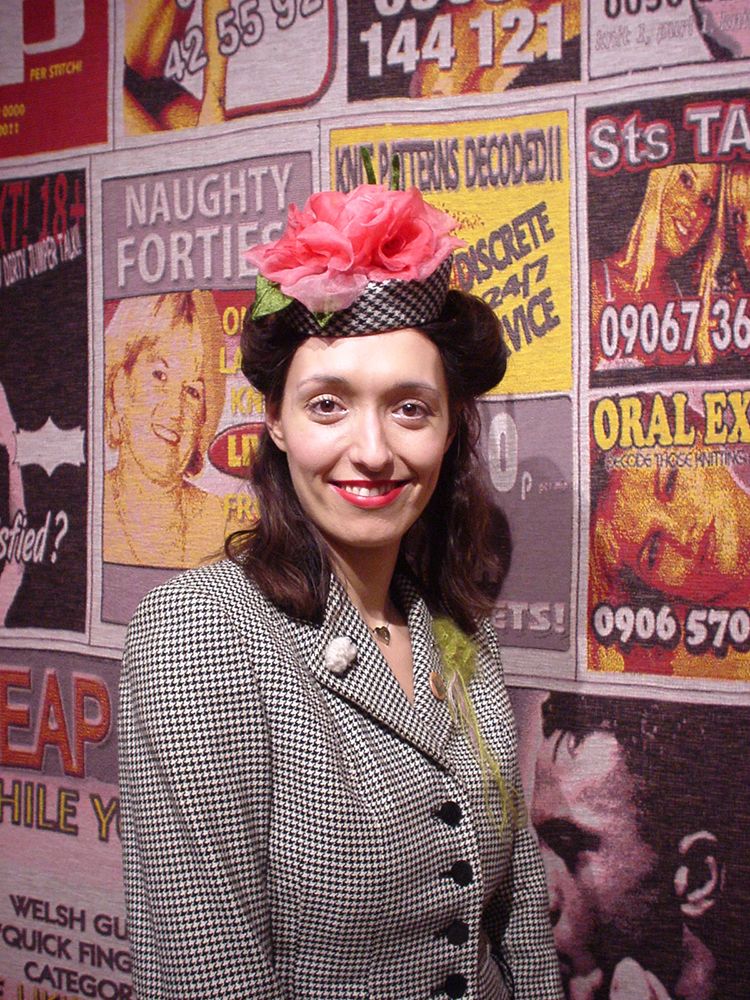 Girl wearing dog-tooth pattern pillbox hat adorned with large pink flower and green leaf.