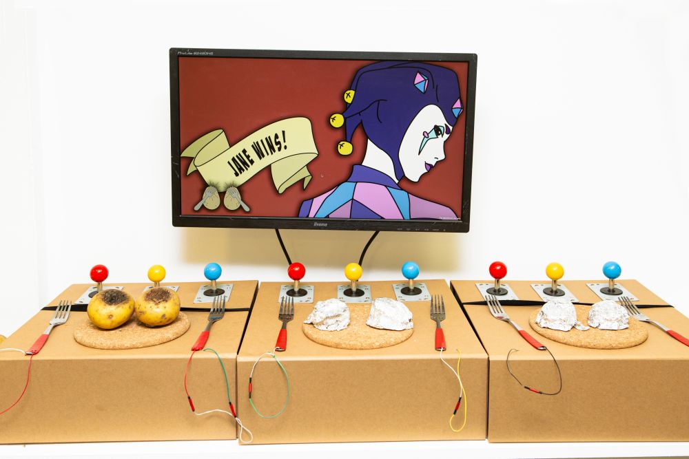 A set-up of games controllers including a knife and fork and wired-up jacket potatoes, all in front of a screen.