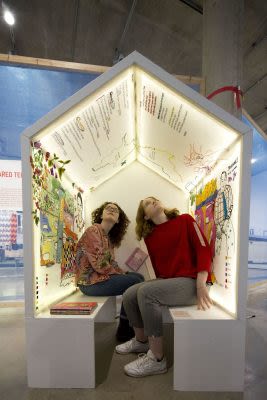 Photograph of a white shed with the from end open and two benches inside. Two people are sat on the benches looking at images and writing on the inside walls of the shed