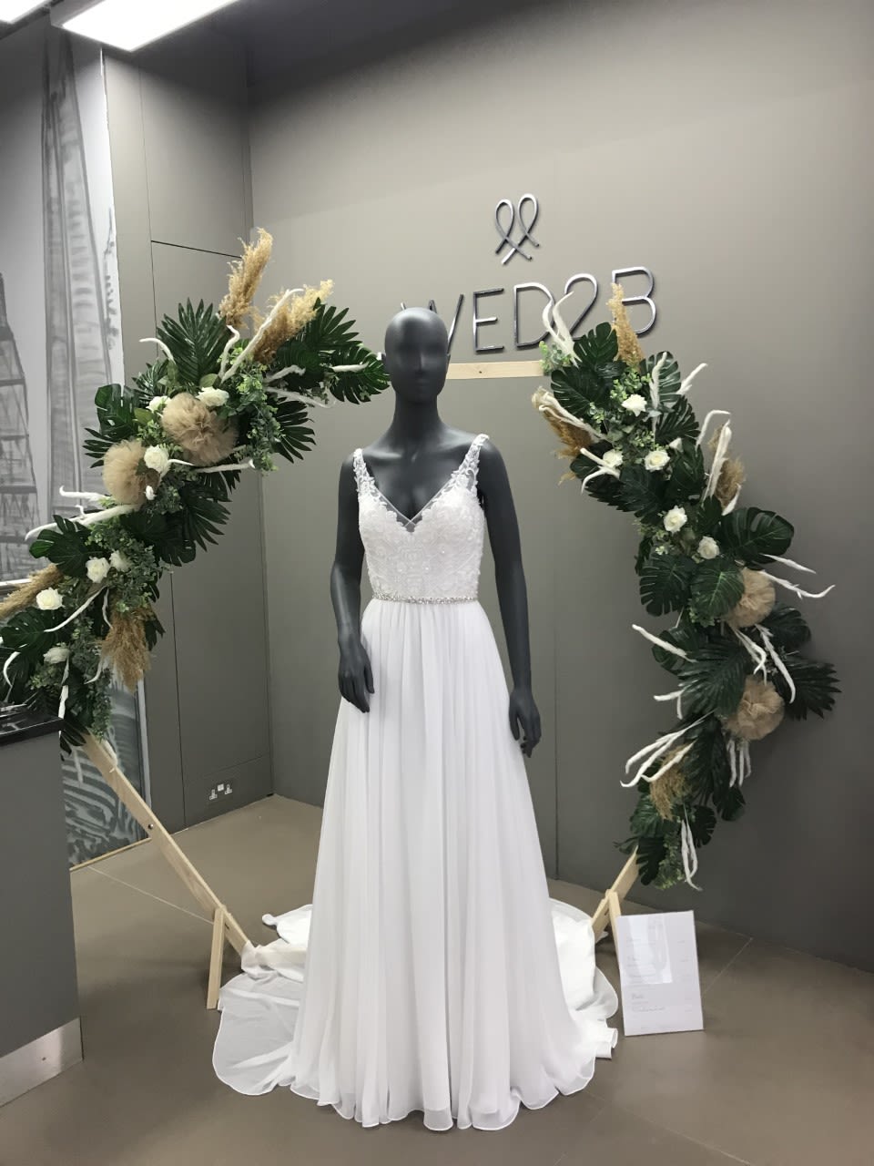 A photo of a mannequin wearing a long white dress with the logo WED2B on the wall behind.