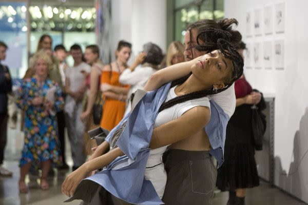 Photograph of two people performing in the gallery while the audience stands watching behind them, one performer is leaning back into the other