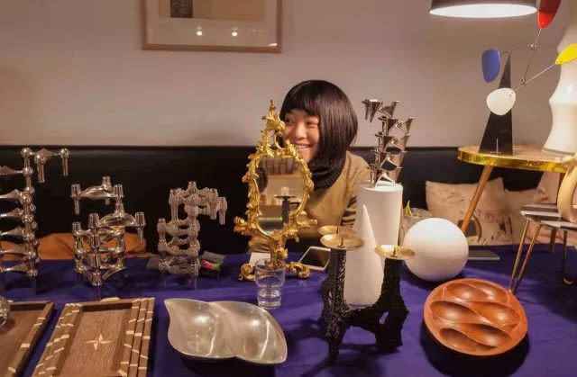 A table with wooden sculptures on in the foreground, and a woman smiling behind