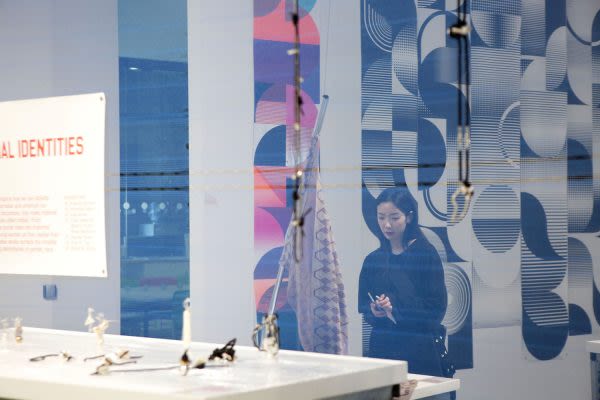 Photograph through a translucent screen of someone looking at artwork displayed on a table, abstract patterns hung on the wall behind them