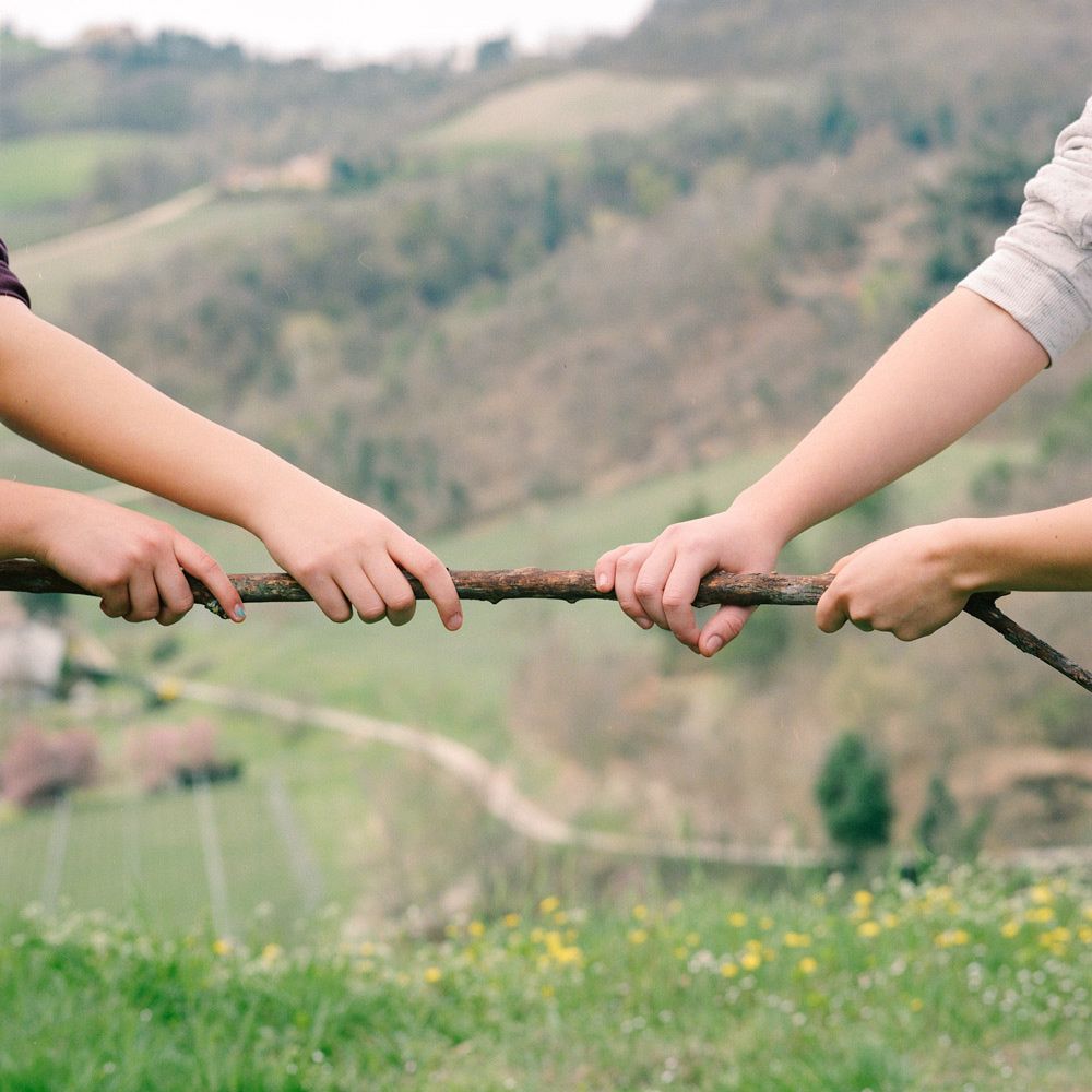 Photo of the arms of two people pulling at a stick in opposing directions, in front of a grassy hill