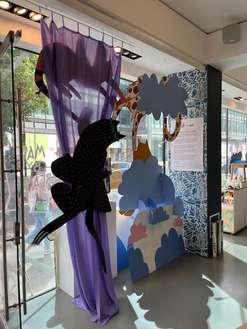 Interior view of a playful and immersive window display at ‘not just a shop’. The scene features layered cut-outs of stylised clouds, a spotted creature resembling a giraffe, and a large black panther figure climbing across a sheer purple curtain. The curtain hangs beside the shop’s glass entrance, allowing daylight to filter through the display. A printed statement about the installation is mounted on a blue and white patterned backdrop on the wall. To the right, part of the shop interior with shelves and colourful products is visible in the background.