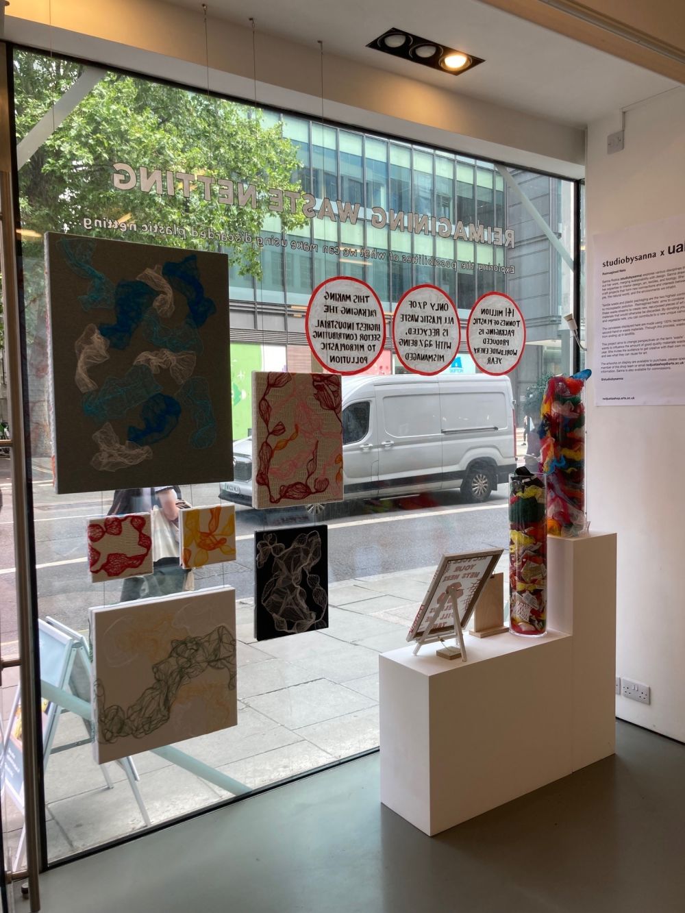 A window display inside 'not just a shop' featuring an installation by studiobysanna. Various abstract textile artworks in different colours are suspended at different heights in front of the shop window, each mounted on square or rectangular canvas. On the window, vinyl text reads “REIMAGINING WASTE NETTING,” alongside several red-circled environmental facts about plastic pollution and recycling. Below the hanging artworks, a white plinth displays tall tubes filled with colourful fabric scraps, a framed information board, and smaller display items. The view outside shows a city street with a passing pedestrian and a parked white van.