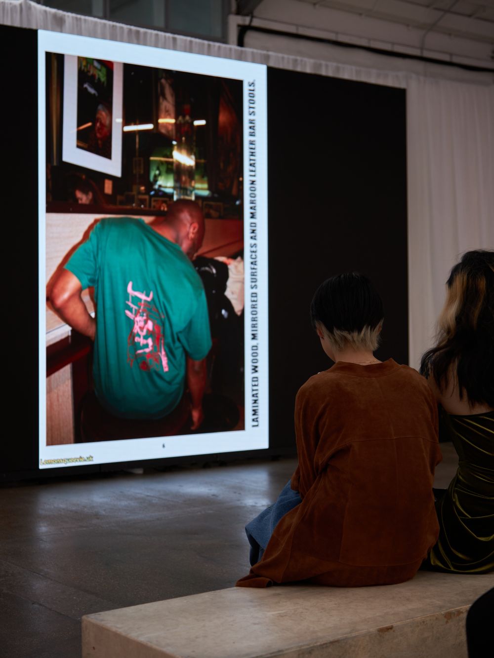 Crowd looking at screen of a person in a green shirt looking down towards the floor
