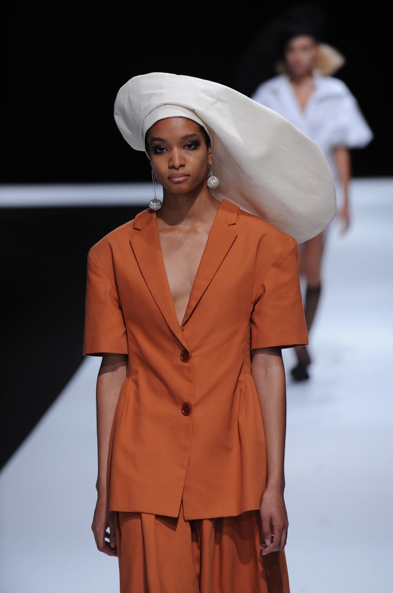 Female model wearing a two piece suit with orange blouse and skirt and big white hat designed by Rachel Simona Cosenza