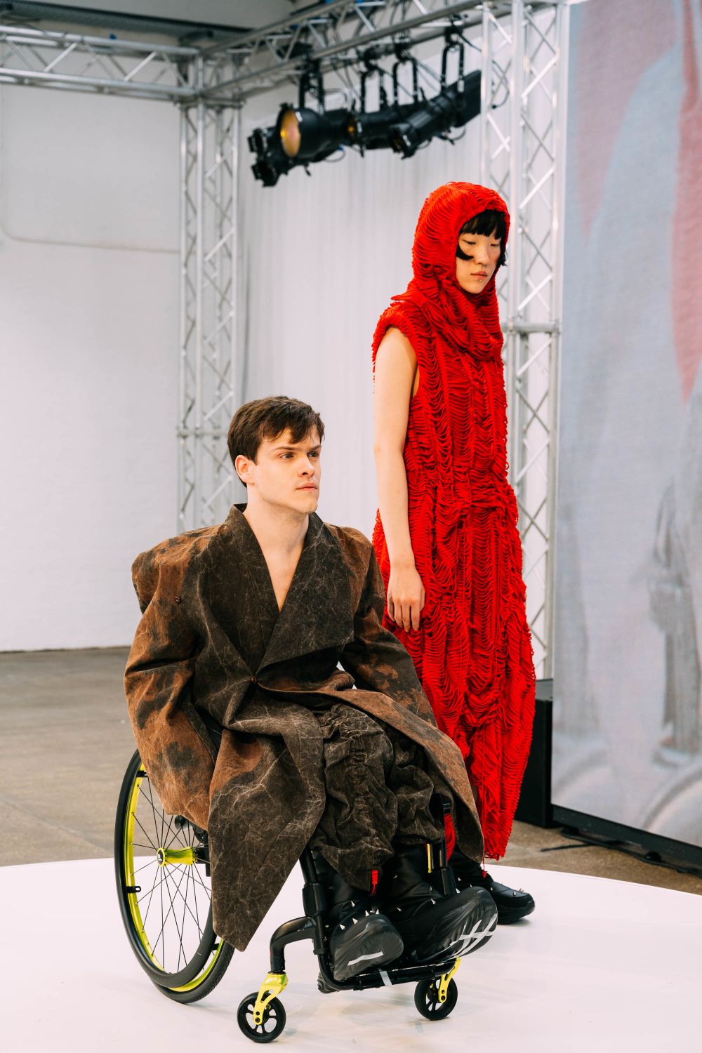 Two models on stage, left using a wheelchair wearing thick brown overcoat, right wearing a red rippled outfit from head to toe