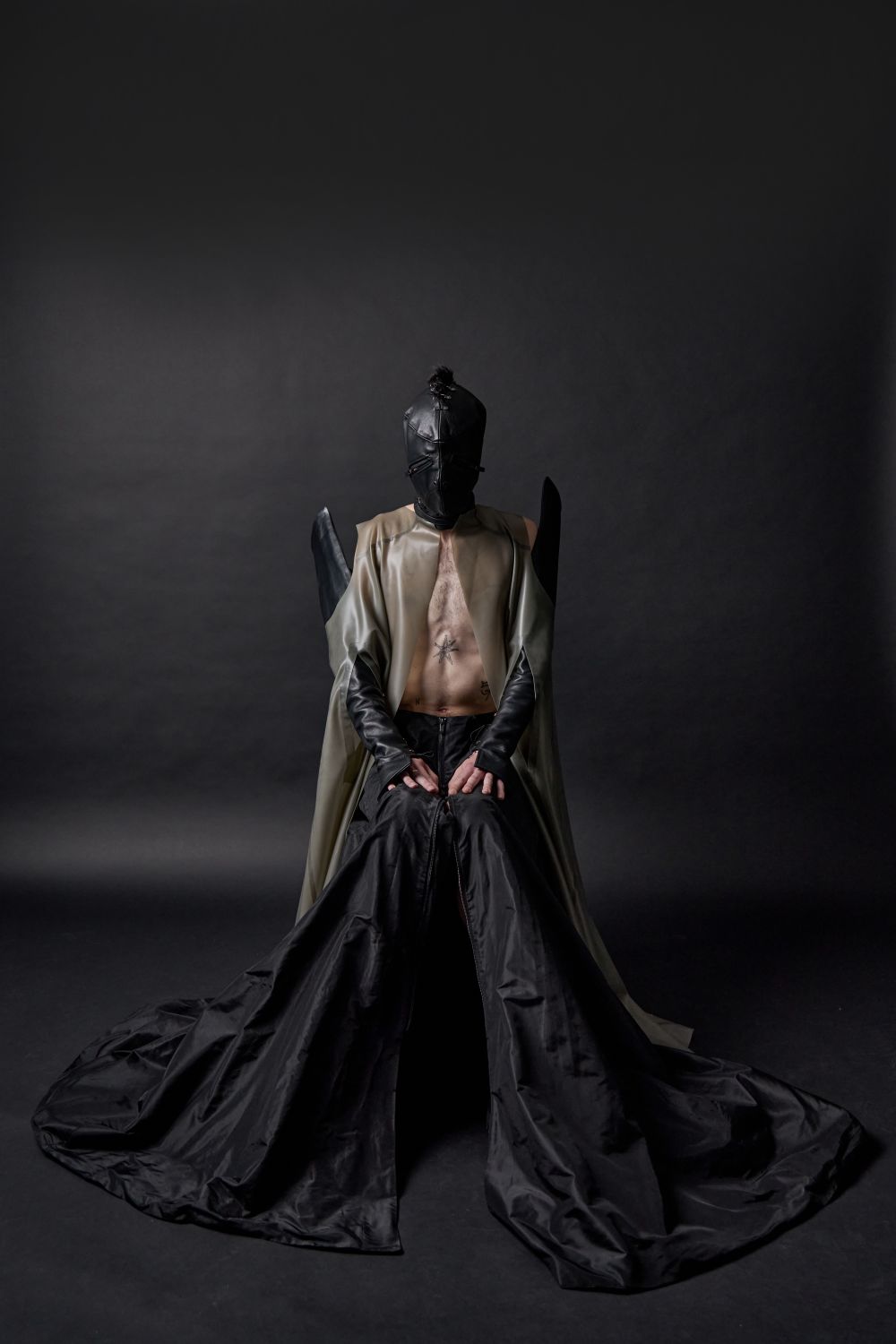 Male model in leather mask, leather cape and wide, trailing black trousers.