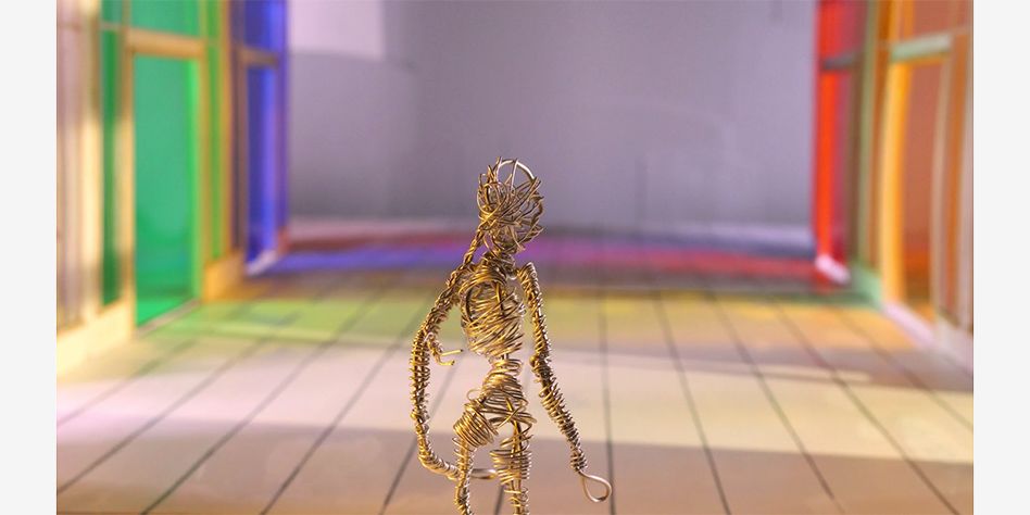 film still of human figure made out of metal wire whilst standing in multicoloured shopping centre