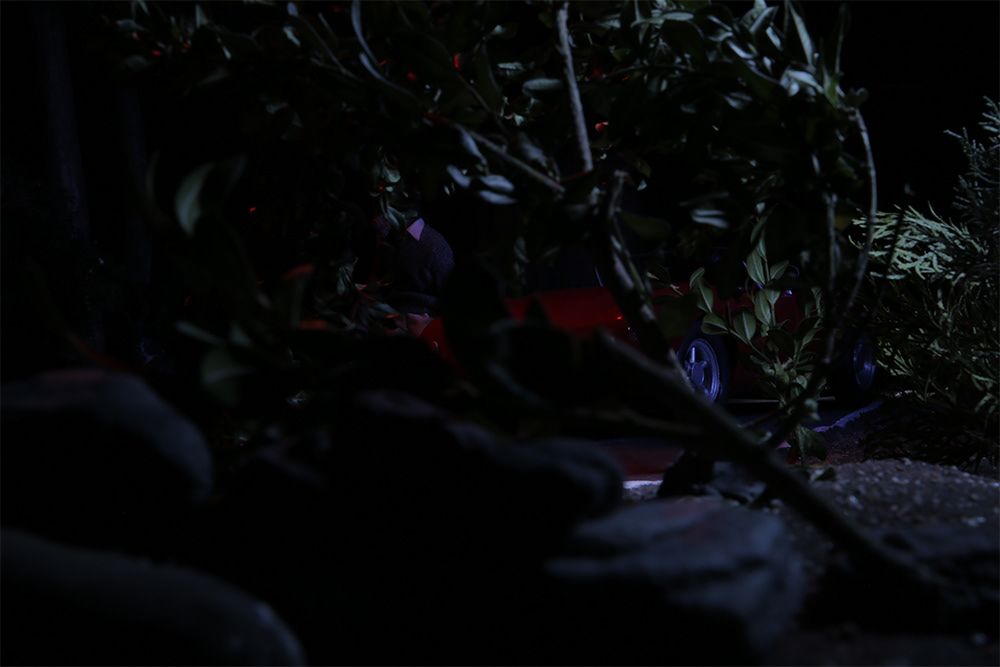 A model car, taken as a still from a stop motion animated film, has crashed into some bushes.