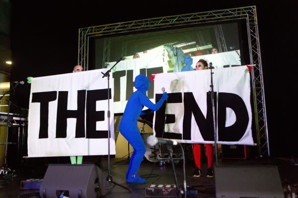 Photograph of two people on a stage holding up signs that say The and End with a third person bent over between them, dressed in a blue morph suit, with a microphone in their hand 
