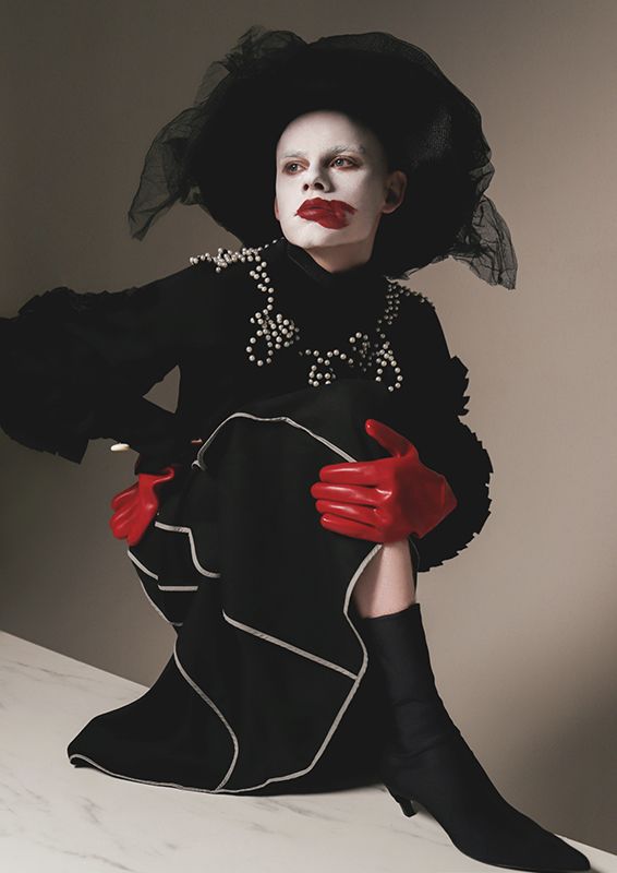 Model wearing a black hat and coat and red gloves with pale white makeup and smudged red lips