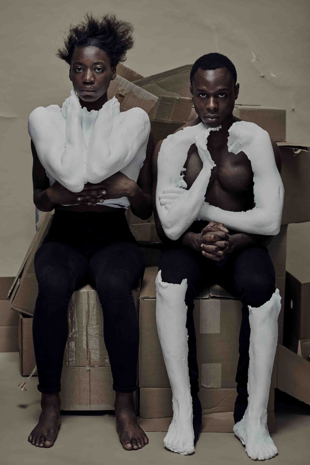 Two models sit on cardboard boxes while wearing plaster-cast items.