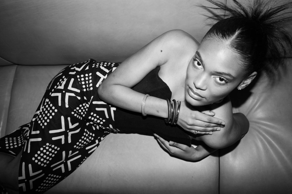 A person lying on a cushioned bench, wearing a patterned skirt and a strapless top, with bracelets on one arm in a black‑and‑white photograph.