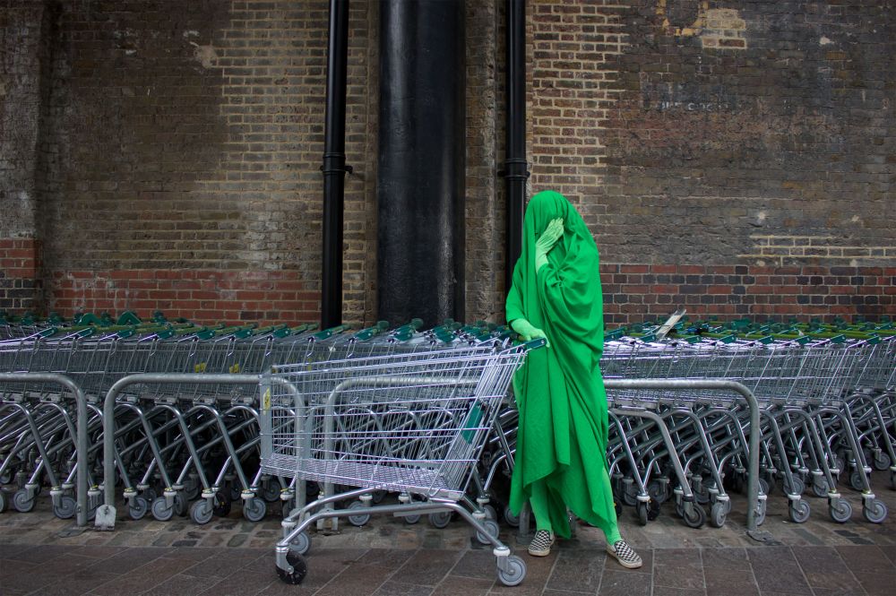 A person dressed entirely in bright green stands next to a row of shopping trolleys 